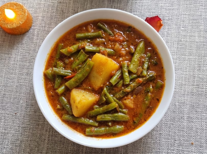 A white bowl of green bean and potato curry, with a lit candle and rose petal nearby.